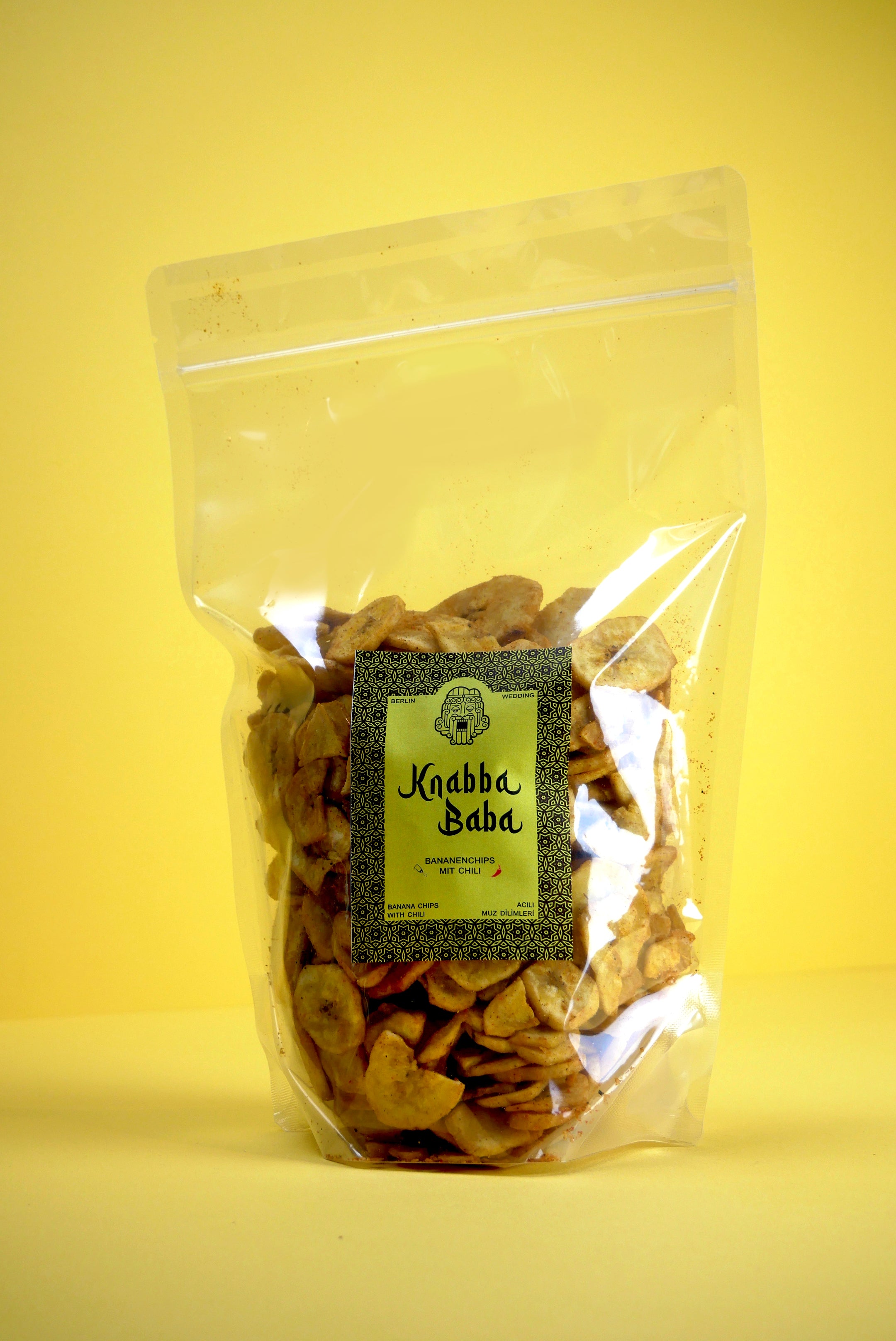 Knabba Baba getrocknete Bananenchips mit Chili 500 g in wiederverschließbarer transparenten Verpackung vor gelbem Hintergrund