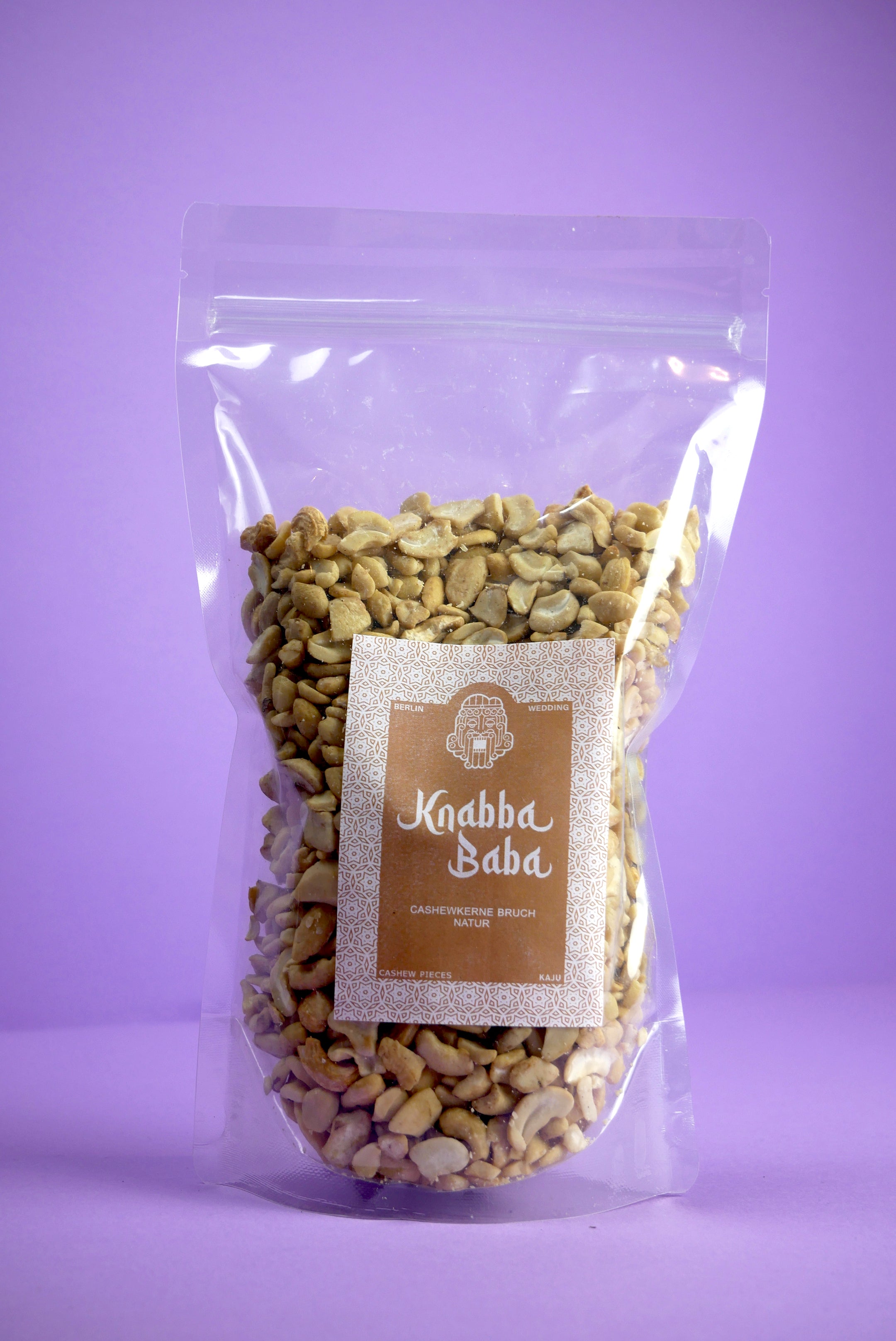 Knabba Baba Cashew Bruch Natur 1000 g im transparenten wiederverschließbaren Beutel vor lila Hintergrund
