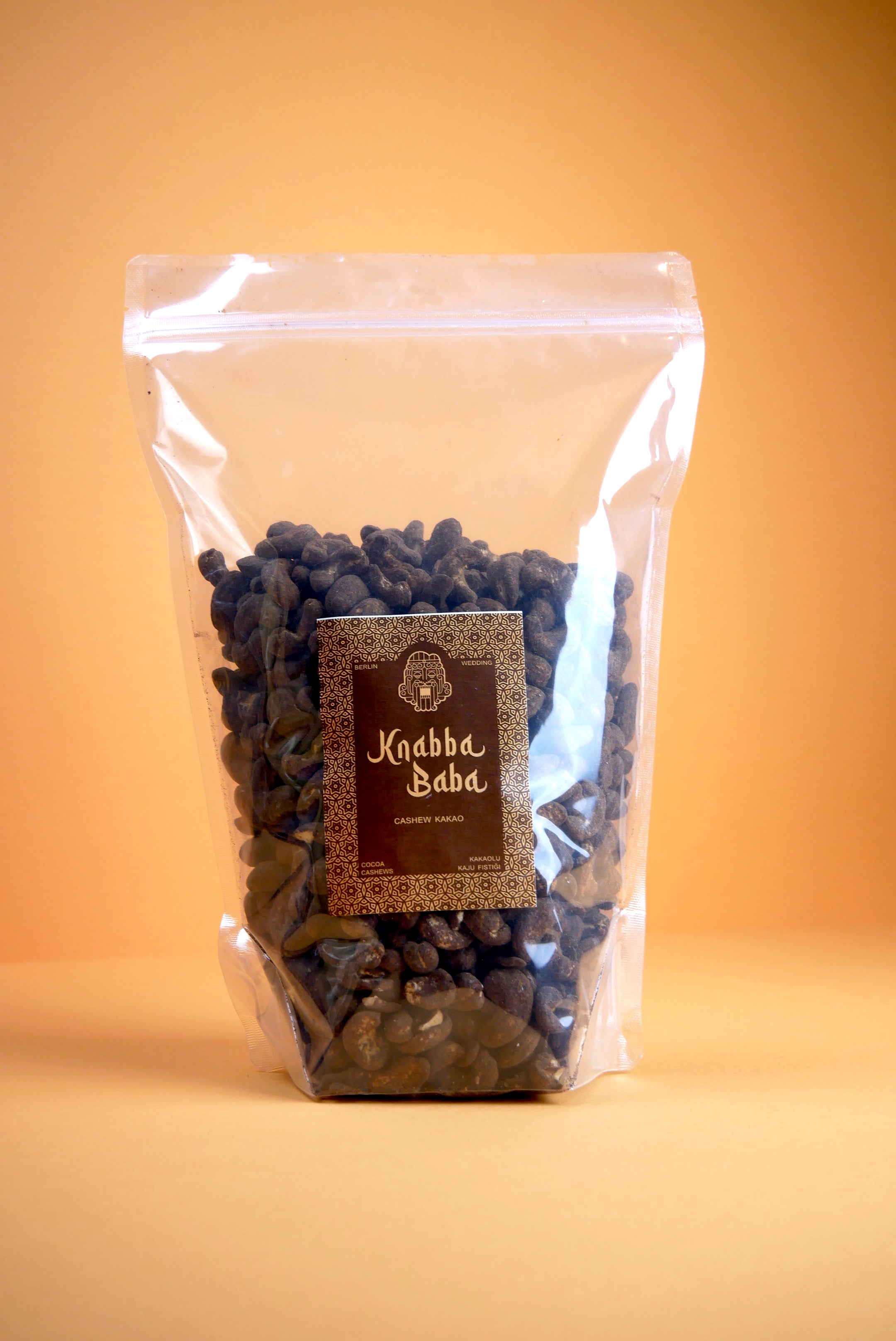 Knabba Baba Cashewkerne mit Kakao 1 kg in durchsichtiger wiederverschließbarer Verpackung mit braunem Etikett auf beigem Hintergrund