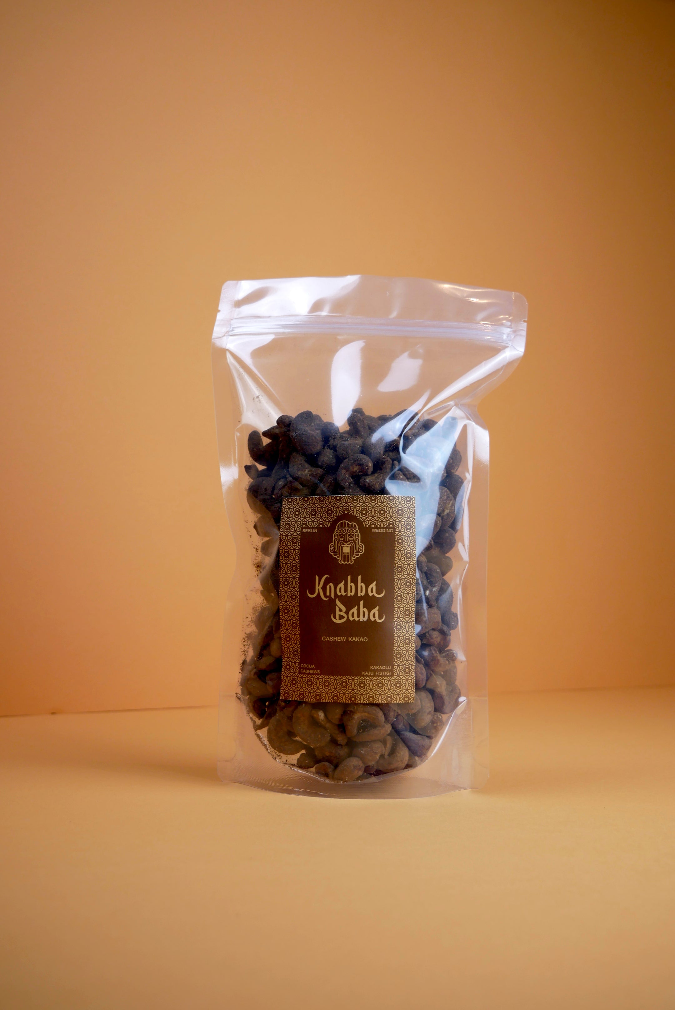 Knabba Baba Cashewkerne mit Kakao 500 g im wiederverschließbaren transparenten Beutel auf beigem Hintergrund