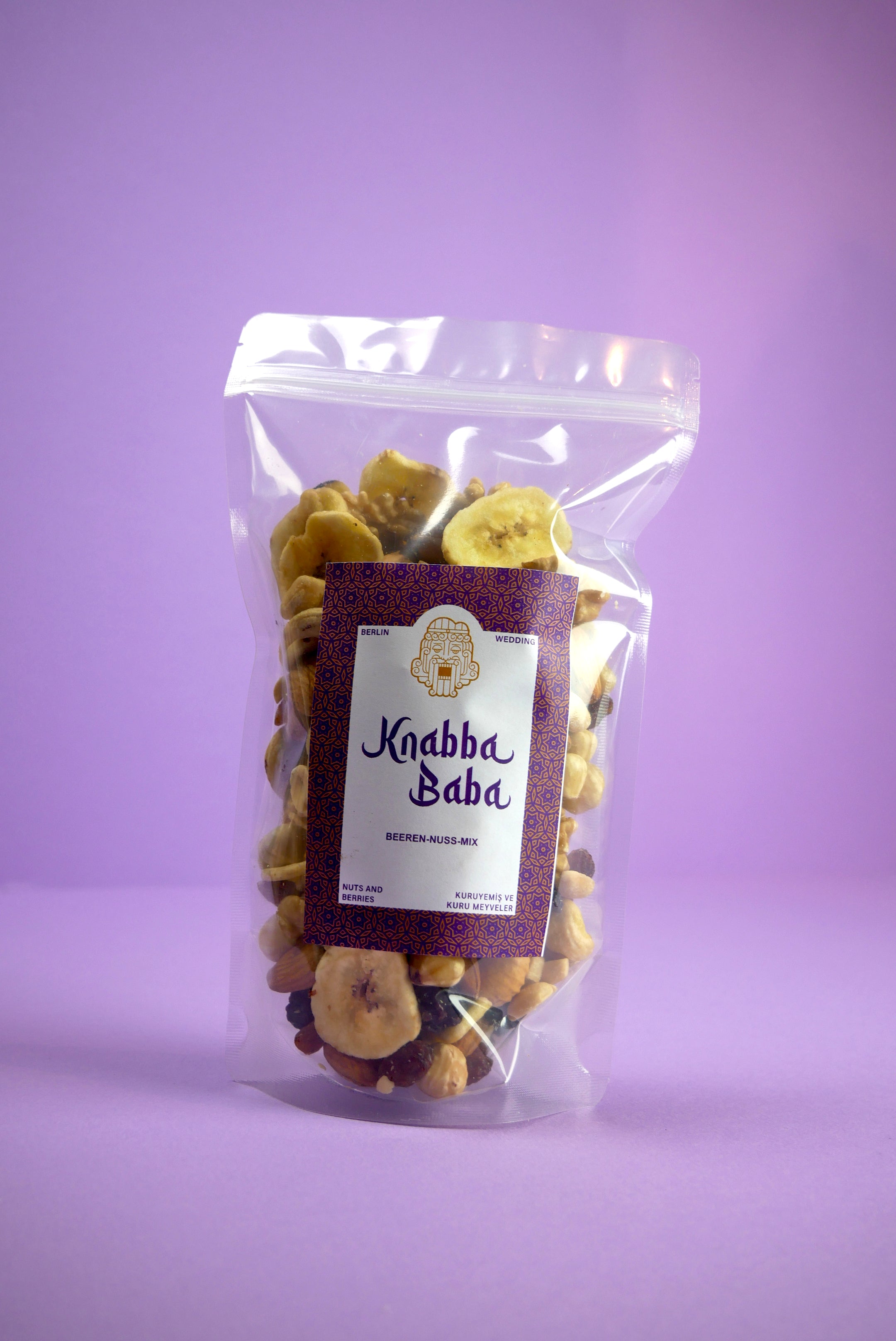 Knabba Baba Nussmischung mit Beeren 250 g  im wiederverschließbaren Beutel mit Bananenchips vor lila Hintergrund
