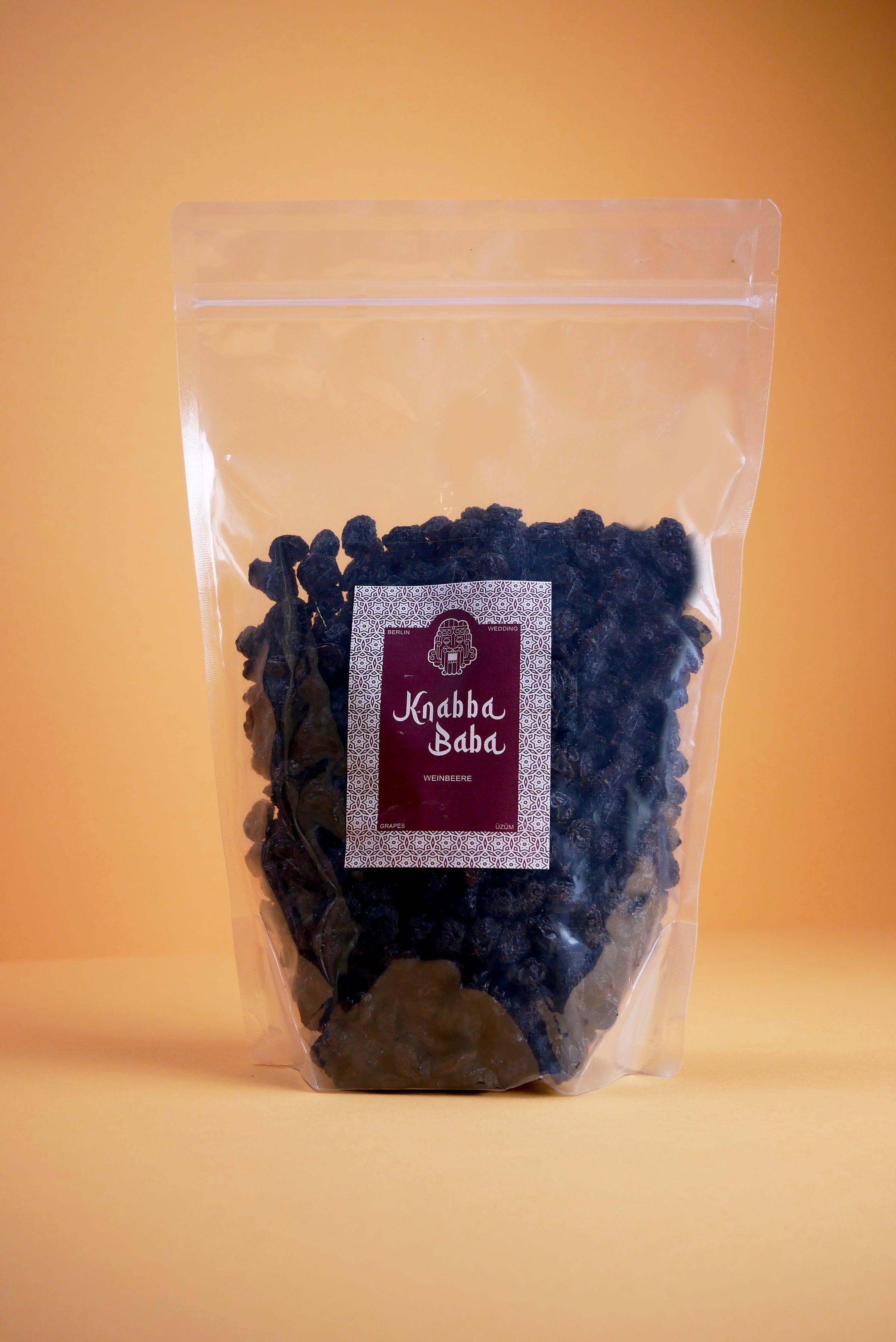 Knabba Baba Weinbeeren (Rosinen) 1 kg in transparentem wiederverschließbarem Beutel vor orangefarbenem Hintergrund